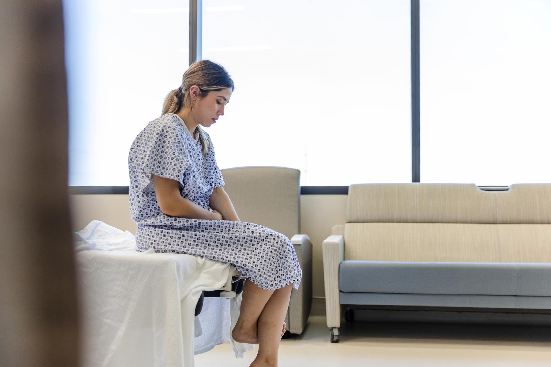 Sad Baton Rouge woman in a hospital gown sitting on the edge of a hospital bed.