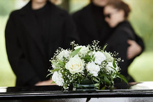 Mourners gather in the background of a casket topped with a bouquet of white flowers.