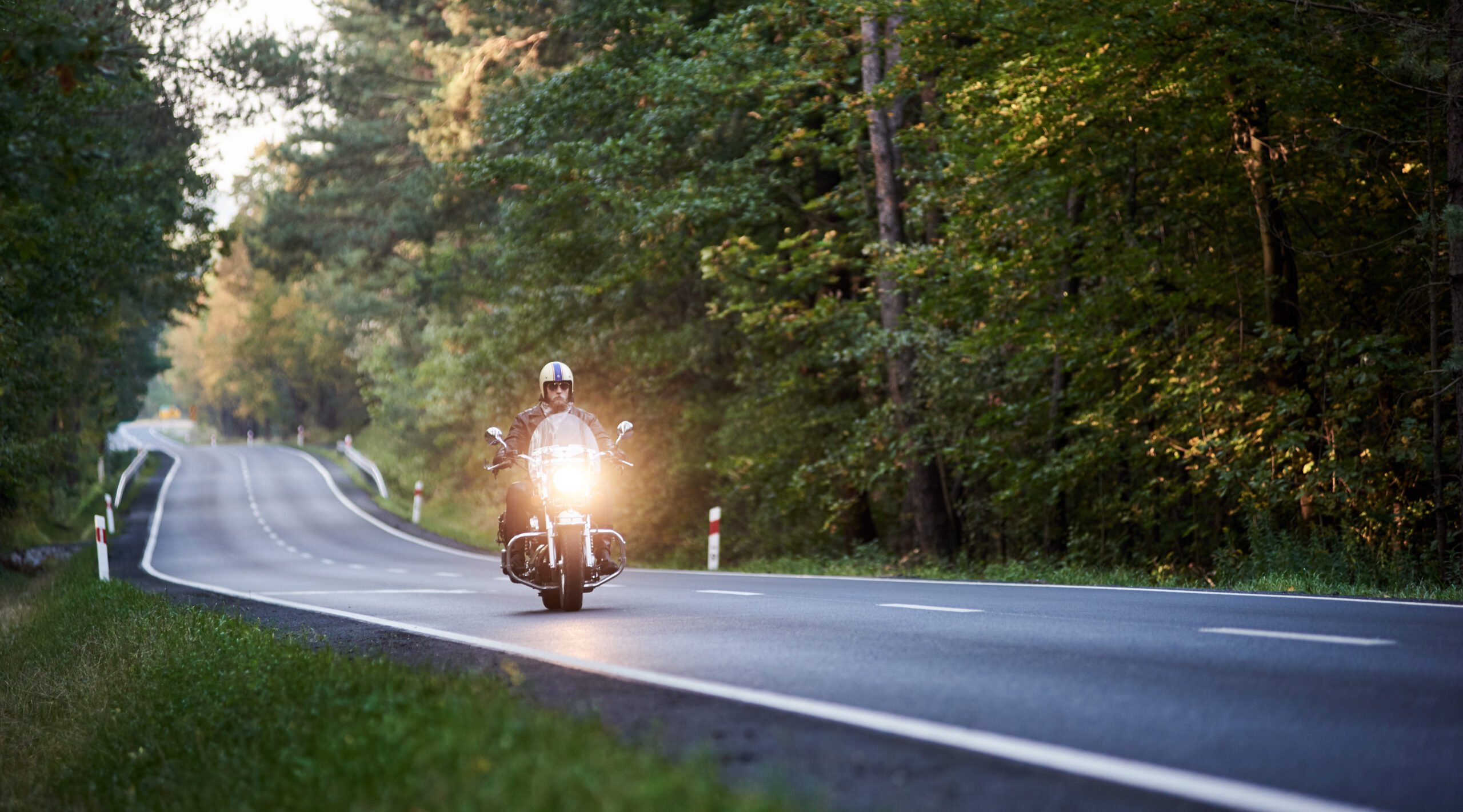 Man wearing a helmet and sunglasses riding a motorcycle down a road surrounded by trees.